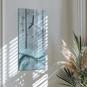 Op de afbeelding: Een rechthoekige wandklok met een lichtblauw houten plankontwerp. De wijzerplaat van de klok heeft zwarte cijfers en wijzers. De onderkant van de klok heeft een blauw-wit golfontwerp. De klok is aan een witte muur bevestigd.