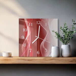 May include: Square wall clock with a red and white abstract design. The clock face features white numerals and hands, with a flowing, wavy pattern in shades of red and white. The clock is displayed on a wooden shelf with decorative items.