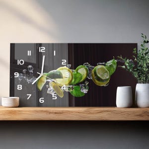 Può includere: Un orologio da parete rettangolare con una stampa grafica di fette di lime, foglie di menta verde fresca e cubetti di ghiaccio trasparenti su sfondo scuro. Il quadrante mostra numeri bianchi da 1 a 12 e due lancette bianche per l'ora.