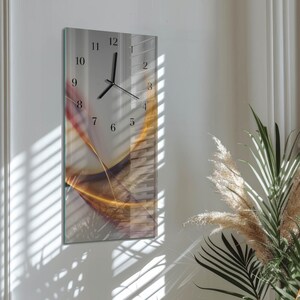 Op de afbeelding: Een rechthoekige wandklok met een glazen voorkant. De klok heeft zwarte wijzers en cijfers tegen een lichte achtergrond met een abstract ontwerp in goud, rood en bruin. De klok is aan een witte muur bevestigd.