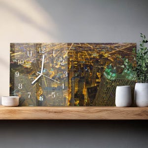 May include: Rectangular wall clock featuring a nighttime cityscape view with illuminated buildings and streets. The clock face displays white numerals and hands. The clock is mounted on a wooden shelf with decorative plants.
