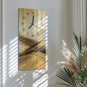 Op de afbeelding: Een rechthoekige wandklok met een beige en bruin abstract ontwerp. De wijzerplaat heeft zwarte cijfers en wijzers. De klok is bevestigd aan een witte muur, met schaduwen van jaloezieën zichtbaar.