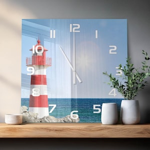 Könnte beinhalten: Eine quadratische Wanduhr mit einem aufgedruckten Bild eines rot-weiß gestreiften Leuchtturms an einer felsigen Küste am Meer. Das Zifferblatt zeigt große weiße Zahlen von eins bis zwölf und zwei dünne weiße Zeiger vor einem blauen Himmel.