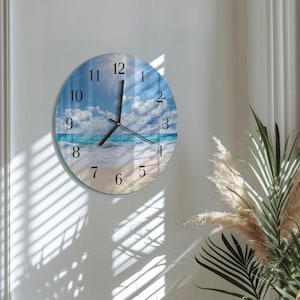 May include: Round wall clock featuring a beach scene with blue water, white clouds, and a sandy shore. The clock has black hands and numerals, with the numbers 1 through 12. The clock is mounted on a white wall.