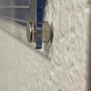 Puede incluir: Primer plano de un letrero de acr&iacute;lico transparente con detalles azules, montado en una pared blanca texturizada. El letrero se sujeta con un separador de metal plateado. El letrero parece tener texto blanco.