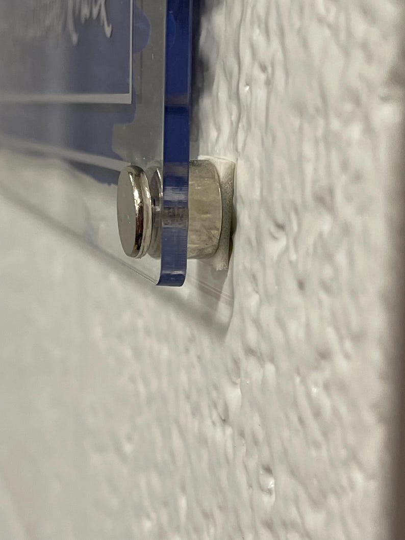 Puede incluir: Primer plano de un letrero de acr&iacute;lico transparente con detalles azules, montado en una pared blanca texturizada. El letrero se sujeta con un separador de metal plateado. El letrero parece tener texto blanco.