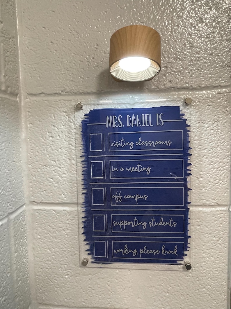 Puede incluir: Un letrero de acr&iacute;lico transparente con el texto "MRS. DANIEL IS" y una lista de verificaci&oacute;n de actividades en letras blancas, montado en una pared de ladrillos blancos. Una l&aacute;mpara de madera y metal est&aacute; encima del letrero.