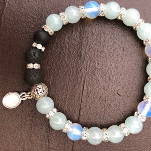 May include: A light blue and white beaded bracelet with a silver charm. The bracelet is made with lava rock beads, aquamarine beads, and opal beads.