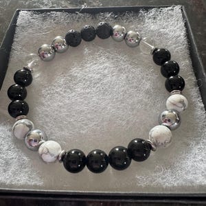 May include: A black, white, and silver beaded bracelet with lava rock beads. The bracelet is made of natural stones and is designed to be worn on the wrist.