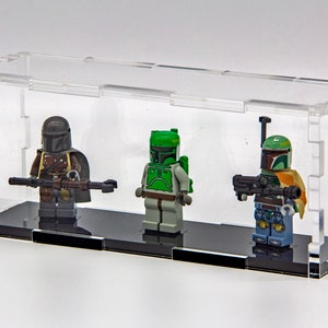 Pode incluir: Três minifiguras Lego Star Wars expostas em uma vitrine de acrílico transparente. As minifiguras são Boba Fett, um Mandaloriano e outro Boba Fett. A vitrine tem uma base preta.
