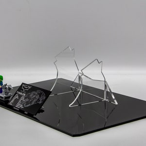 May include: A black and gray Lego Batman and Joker minifigure set on a black base with a clear acrylic display stand. The set includes a black and gray Lego Tumbler vehicle. The base has a black and white card with the text "The Dark Knight Trilogy Tumbler" and a silhouette of the Tumbler.