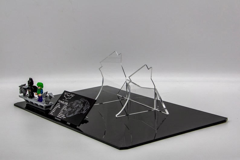 May include: A black and gray Lego Batman and Joker minifigure set on a black base with a clear acrylic display stand. The set includes a black and gray Lego Tumbler vehicle. The base has a black and white card with the text "The Dark Knight Trilogy Tumbler" and a silhouette of the Tumbler.