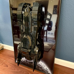 Puede incluir: Un modelo tridimensional enmarcado de una nave espacial, construido con bloques de construcción negros y grises. El modelo está situado sobre un fondo de cielo estrellado y superficie planetaria, con un marco negro.