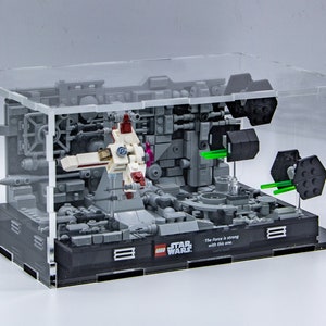 May include: A clear acrylic display case containing a Star Wars Lego set. The set features a grey and black spaceship with green lights, a white and red spaceship, and a grey and black base with the text "Star Wars" and "The Force is strong with this one."