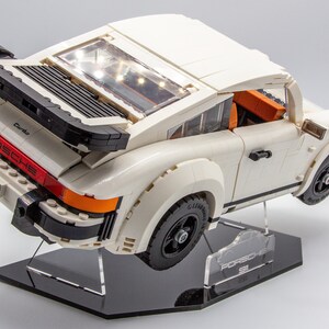 May include: A white Lego Porsche 911 Turbo model with black accents and a red "PORSCHE" logo on the back. The car is on a black display stand with a clear acrylic plate that says "PORSCHE 911".