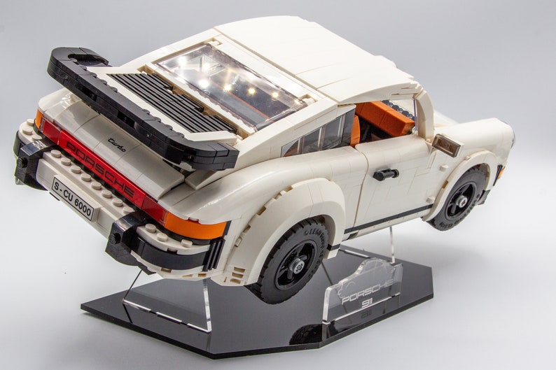 May include: A white Lego Porsche 911 Turbo model with black accents and a red "PORSCHE" logo on the back. The car is on a black display stand with a clear acrylic plate that says "PORSCHE 911".