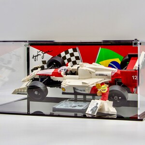 May include: A white, red, and black LEGO Formula 1 race car with a Shell logo and the text "H HONDA" on the side. The car is displayed in a clear acrylic case with a black base. The case also includes a LEGO minifigure of a race car driver wearing a red and white racing suit and a helmet. The background of the case features a checkered flag and a Brazilian flag.
