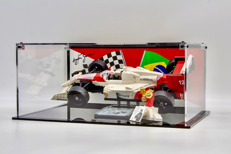 May include: A white, red, and black LEGO Formula 1 race car with a Shell logo and the text "H HONDA" on the side. The car is displayed in a clear acrylic case with a black base. The case also includes a LEGO minifigure of a race car driver wearing a red and white racing suit and a helmet. The background of the case features a checkered flag and a Brazilian flag.