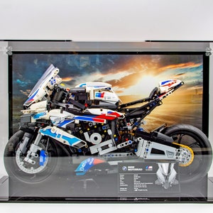 Könnte beinhalten: Ein weißes, blaues und rotes LEGO® Technic™ BMW M 1000 RR Motorradmodell in einer transparenten Vitrine mit schwarzem Sockel. Das Motorrad steht auf einem schwarzen Sockel mit einem gedruckten Hintergrund eines Sonnenuntergangs und einer Straße. Die Vitrine hat einen schwarzen Sockel und transparente Acrylseiten. Das Motorrad hat ein detailliertes Design mit funktionierender Federung und einem detaillierten Motor. Das Motorrad ist eine Nachbildung des echten BMW M 1000 RR Motorrads.