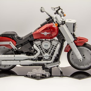 Op de afbeelding: Een rood en zilver LEGO Harley-Davidson motorfietsmodel met een gedetailleerde motor en chromen uitlaatpijpen. De motorfiets staat op een zwarte displaystandaard met een transparante acrylplaat waarop "10285 Harley-Davidson Fat Boy" staat.