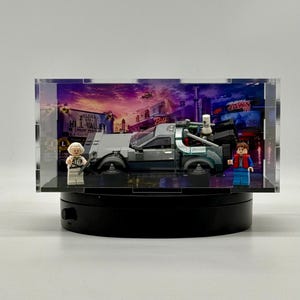 May include: A LEGO Back to the Future DeLorean time machine model displayed inside a clear acrylic case. The set includes two minifigures, one with white hair and one with brown hair, positioned beside the silver car on a black base.