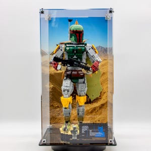 May include: A buildable Boba Fett figure constructed from plastic bricks, displayed inside a clear rectangular acrylic case. The figure features a green helmet, chest armour, yellow knee pads, and a blaster rifle. A small information plaque is at the base.