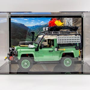 May include: A green and white toy SUV, with a black roof rack and spare tyre, displayed in a clear acrylic case. The SUV has a detailed design, with a mountain backdrop. The case is on a black base.