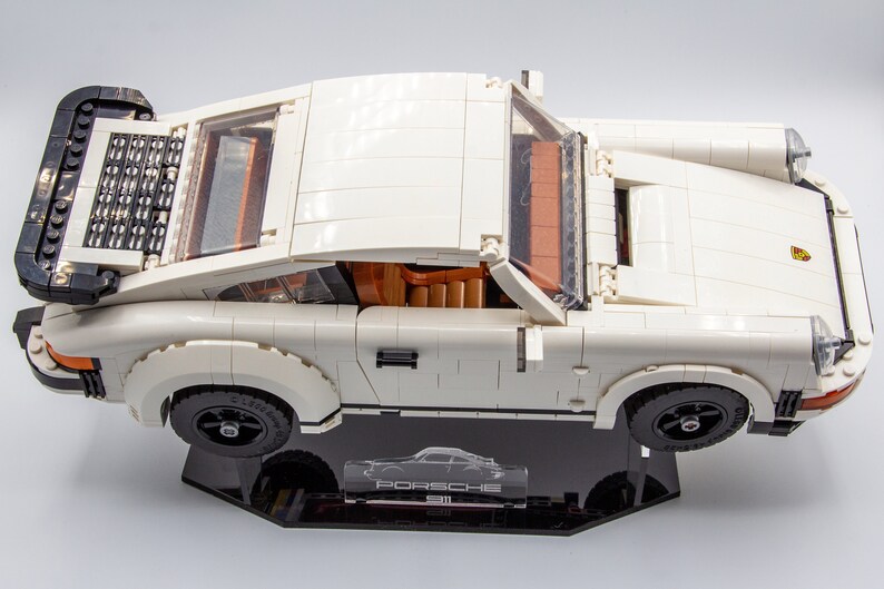 May include: A white Lego Porsche 911 Turbo model with black accents and a clear windscreen. The model is displayed on a black display stand with the word "Porsche" printed on it.