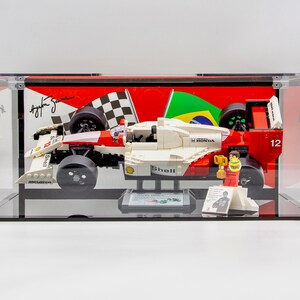May include: A red, white, and black LEGO Formula 1 race car model in a clear display case. The car has the number 12 on the side and the words "Shell" and "Honda" on the body. The car is on a black base with a small LEGO figure of a race car driver holding a trophy.