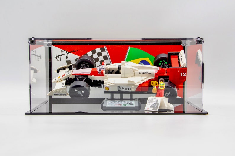 May include: A red, white, and black LEGO Formula 1 race car model in a clear display case. The car has the number 12 on the side and the words "Shell" and "Honda" on the body. The car is on a black base with a small LEGO figure of a race car driver holding a trophy.
