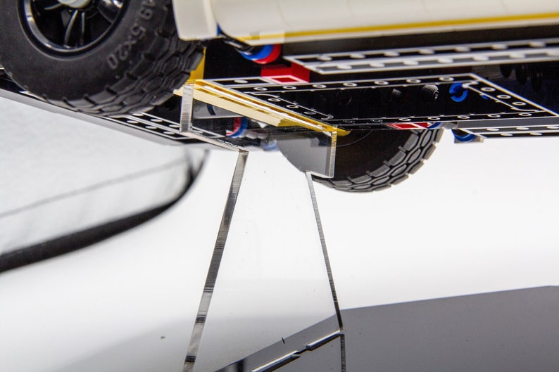 May include: A white Lego vehicle with black tyres and a yellow stripe on the bottom. The vehicle is on a clear acrylic display stand.