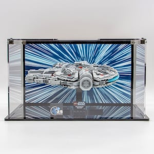 May include: A clear acrylic display case with a black base and a black background. Inside the case is a LEGO Star Wars Millennium Falcon model, with a blue and white light speed effect behind it. The case has a small plaque on the bottom that says "LEGO Star Wars 25th Anniversary".