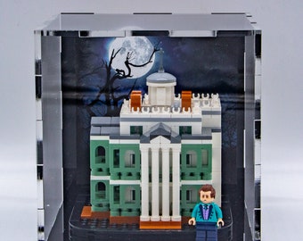 May include: A clear acrylic display case containing a green and white Lego model of a haunted mansion. The model is set against a dark background with a moon and a tree. A small Lego figure in a green jacket and blue trousers stands in front of the mansion.