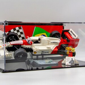 May include: A white and red Lego Formula 1 race car with a Shell logo and Honda engine, displayed in a clear acrylic case with a checkered flag and Brazilian flag backdrop. The car is signed by Ayrton Senna.