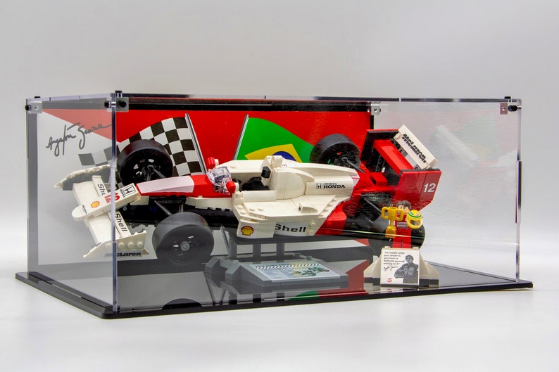 May include: A white and red Lego Formula 1 race car with a Shell logo and Honda engine, displayed in a clear acrylic case with a checkered flag and Brazilian flag backdrop. The car is signed by Ayrton Senna.