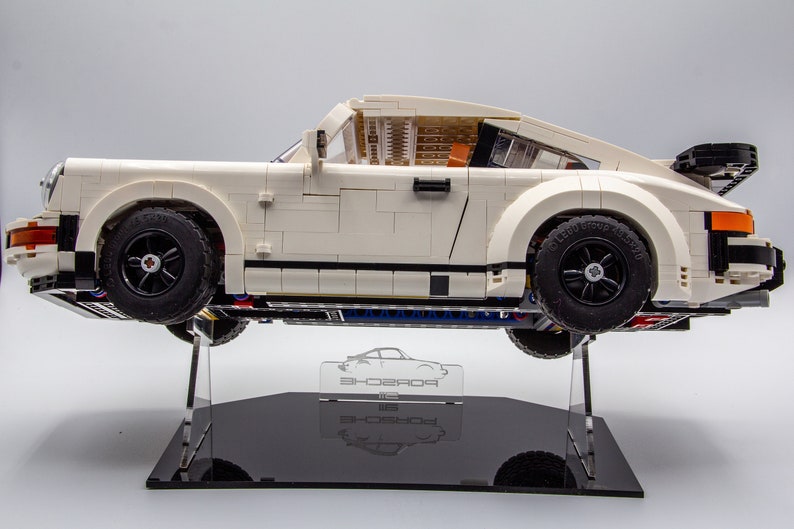 May include: A white Lego Porsche 911 Turbo model on a black display stand. The car is on a clear acrylic stand with a Porsche logo.