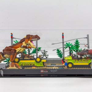 May include: A brown Tyrannosaurus Rex dinosaur Lego set in a clear display case. The dinosaur is attacking a green Jeep with a figure inside. The set includes palm trees and a fence.
