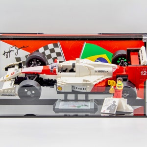 May include: A white, red, and black Lego model of a Formula 1 race car with the number 12 on the side. The car is in a clear display case with a black base. The car is on a black base with a small figure of a race car driver holding a trophy. The background of the display case is a red and white checkered flag, a green and yellow Brazilian flag, and a signature.