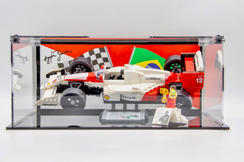 May include: A white, red, and black Lego model of a Formula 1 race car with the number 12 on the side. The car is in a clear display case with a black base. The car is on a black base with a small figure of a race car driver holding a trophy. The background of the display case is a red and white checkered flag, a green and yellow Brazilian flag, and a signature.