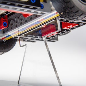 May include: A close-up of the underside of a toy truck, showing the black and grey plastic chassis, red and blue Lego bricks, and a clear acrylic display stand.