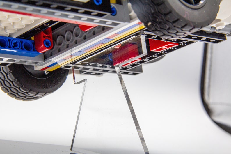 May include: A close-up of the underside of a toy truck, showing the black and grey plastic chassis, red and blue Lego bricks, and a clear acrylic display stand.