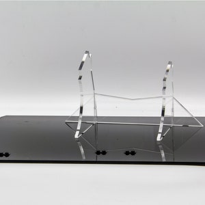 May include: A clear acrylic display stand for a sword or other long object. The stand is made of two pieces that are connected in the middle. The stand is sitting on a black surface.