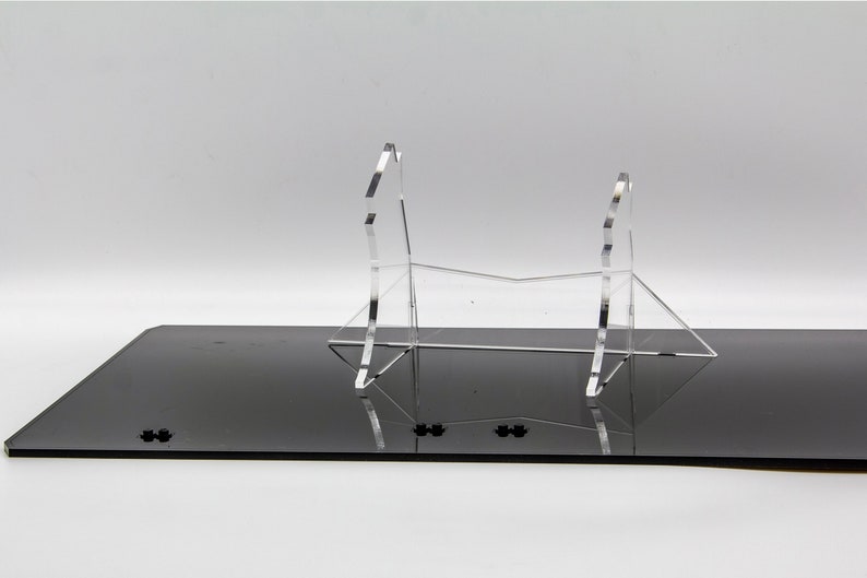 May include: A clear acrylic display stand for a sword or other long object. The stand is made of two pieces that are connected in the middle. The stand is sitting on a black surface.