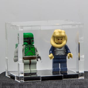 May include: Two Lego minifigures, one in a green and gray Mandalorian armor and helmet, and another with a yellow head and a blue outfit, are displayed inside a clear acrylic case. The case sits on a black base.