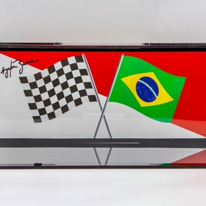 May include: A clear acrylic display case with a black base, showcasing a checkered flag and a Brazilian flag crossed together. The checkered flag is black and white, and the Brazilian flag is green, yellow, and blue with a white circle in the center. The case has a signature on the top, "Ayrton Senna".