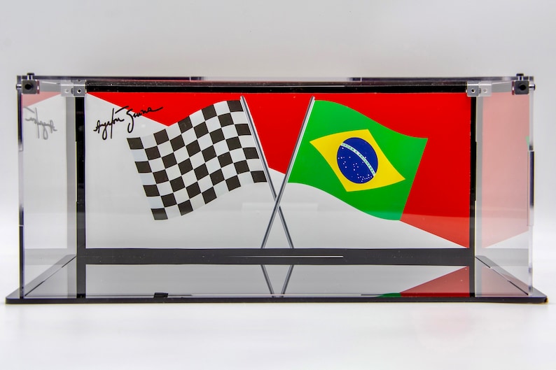 May include: A clear acrylic display case with a black base, showcasing a checkered flag and a Brazilian flag crossed together. The checkered flag is black and white, and the Brazilian flag is green, yellow, and blue with a white circle in the center. The case has a signature on the top, "Ayrton Senna".