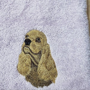 May include: A lavender-colored towel with an embroidered image of a golden brown cocker spaniel dog.