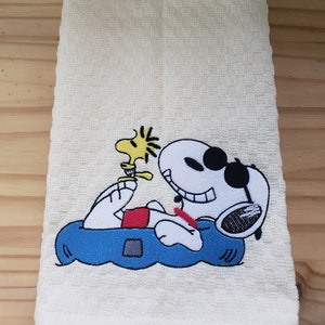 May include: A white kitchen towel with an embroidered image of Snoopy and Woodstock. Snoopy is wearing sunglasses and is floating on a blue inner tube. Woodstock is perched on Snoopy's arm.