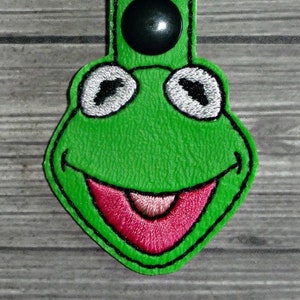 May include: Green felt keychain with an embroidered frog face. The frog has a big smile and large eyes. The keychain has a silver metal ring.