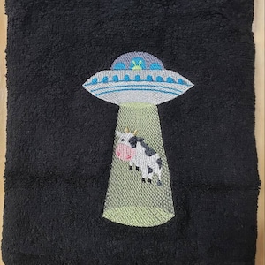 May include: A black towel with an embroidered design of a UFO abducting a cow. The UFO is gray with blue accents and the cow is black and white.